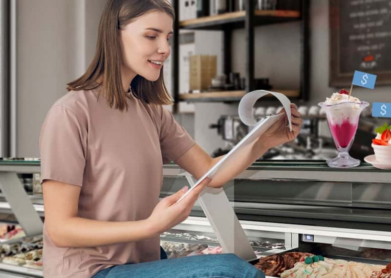 mujer revisando tabla sujetadora de hojas en frente vitrina refrigerada y helados de diferente presentación